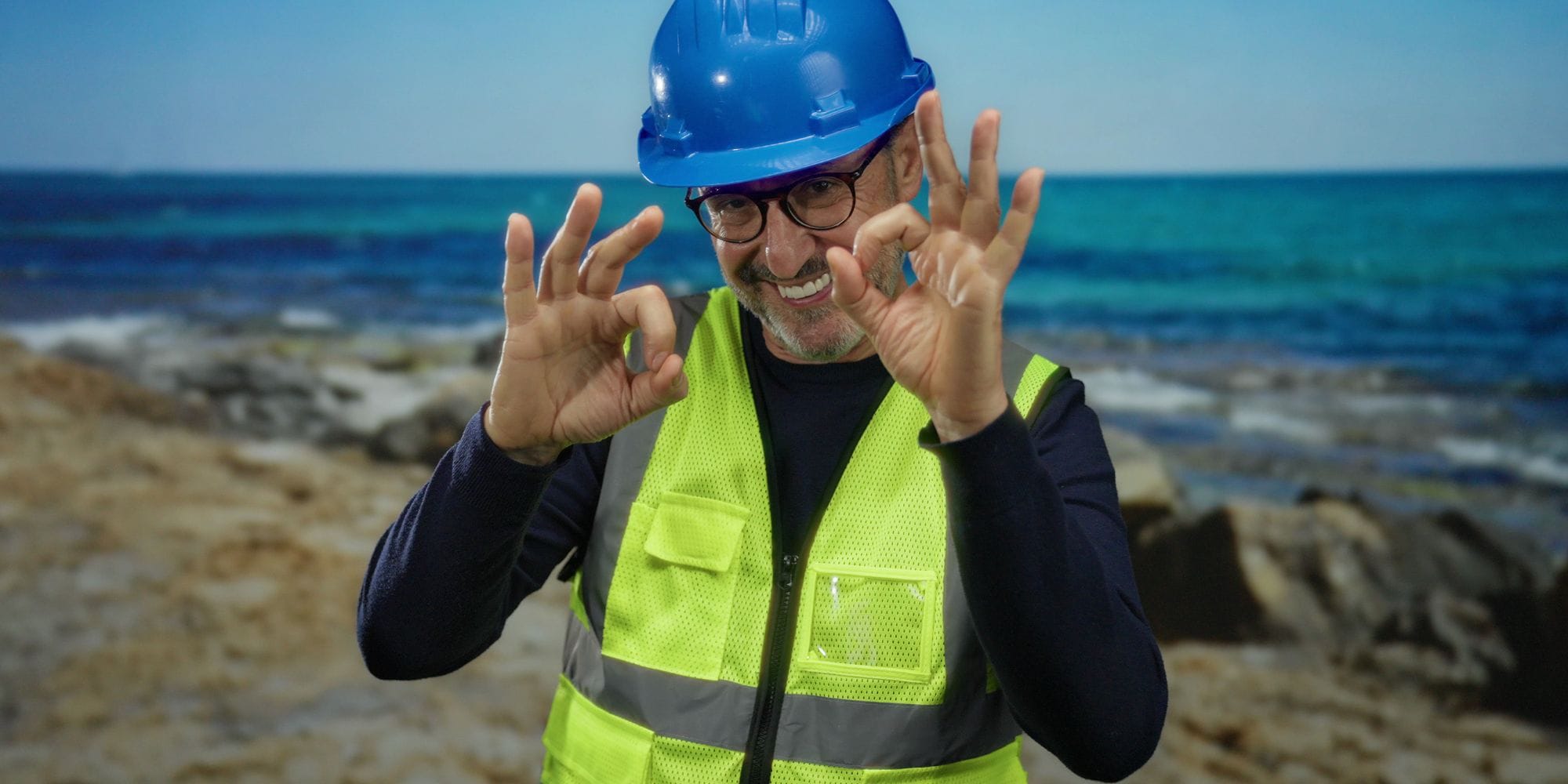 Contractor in a hardhat and safety vest giving the okay sign as he stands at the seacoast